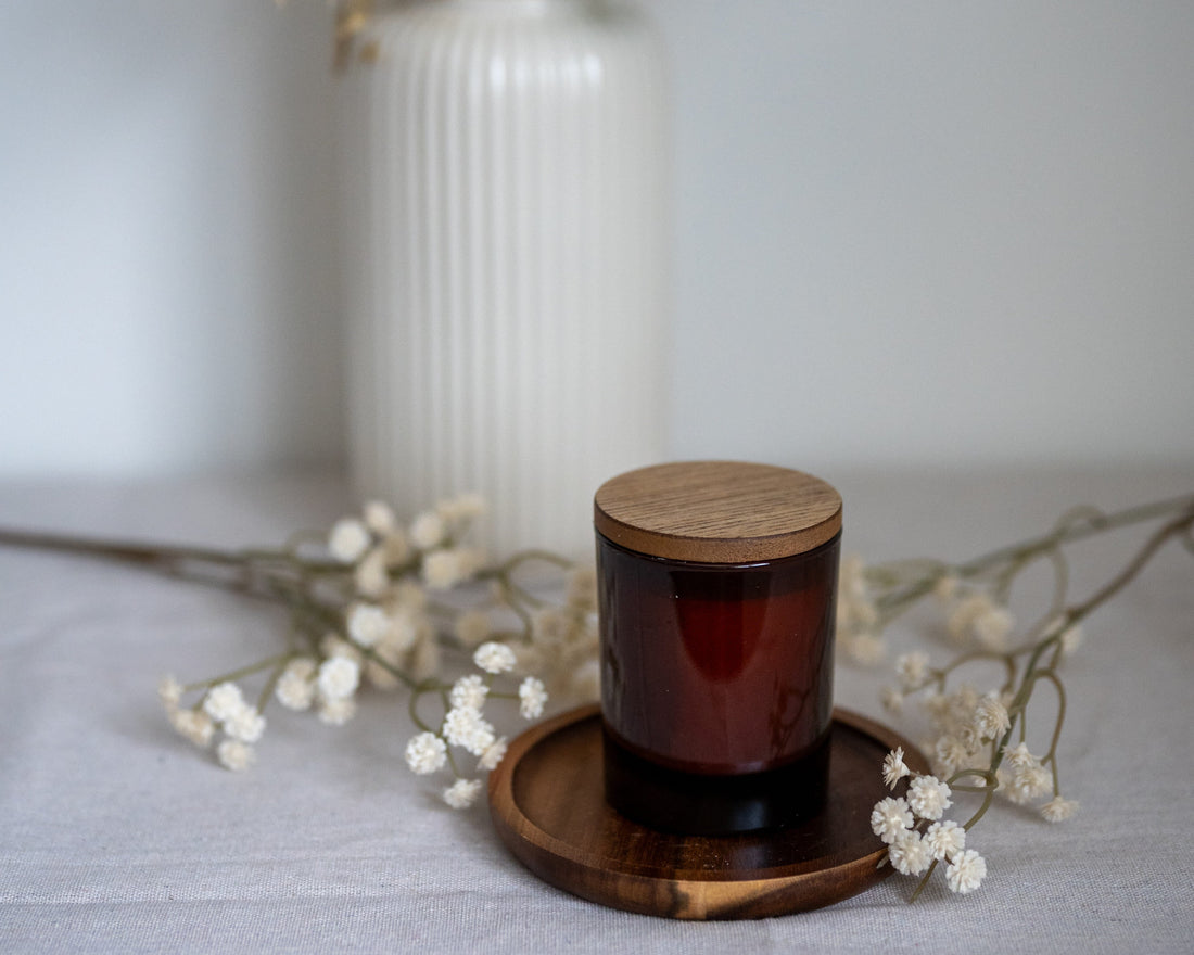 geurkaars in bruin glazen pot met houten deksel op houten onderzetter met witte bloemen op de achtergrond